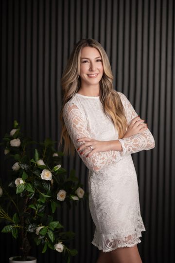 Woman in white lace dress with arms crossed, smiling. Posed in front of a dark, textured wall and flowers.