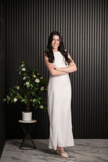 Woman in white dress standing, arms crossed, smiling. Beside a potted plant against a dark, ridged wall.