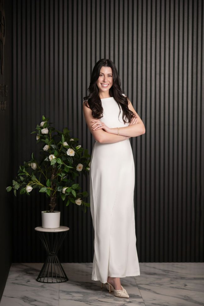 Woman in white dress standing, arms crossed, smiling. Beside a potted plant against a dark, ridged wall.