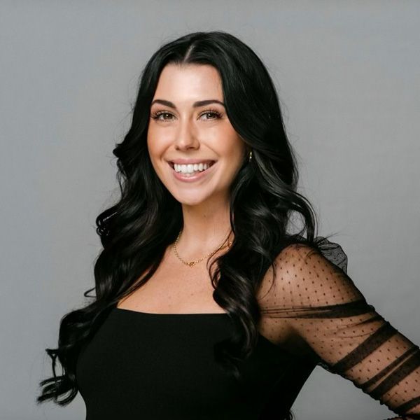 Woman with dark wavy hair smiles, wearing a black top with sheer sleeves, against a gray backdrop.