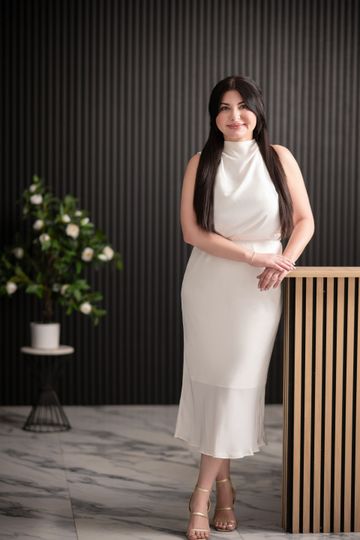 Woman in white dress leans on a wooden counter, smiling. Dark wall behind her, marble floor.