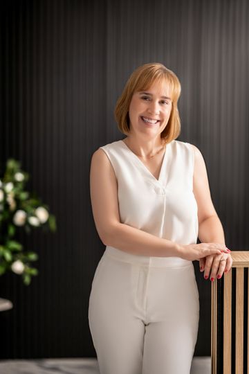 Woman in white outfit smiles, leaning on a wooden counter with black vertical background.