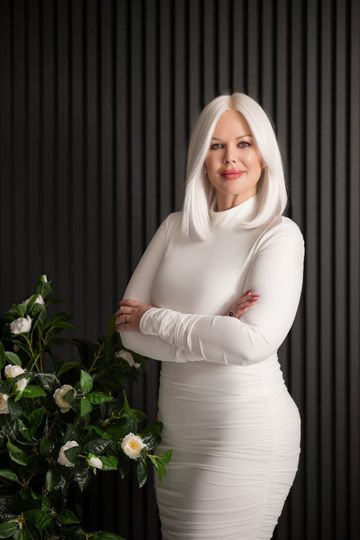 Woman with white hair in white dress, arms crossed, standing near flowers, against a dark background.