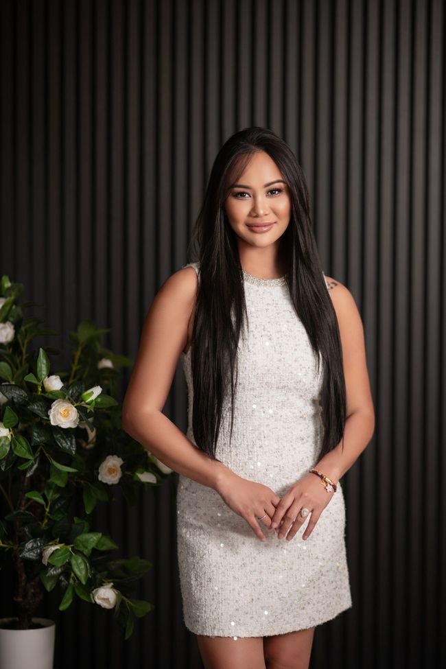 Woman in a white sequined dress stands smiling, hands clasped, near a plant against a dark background.