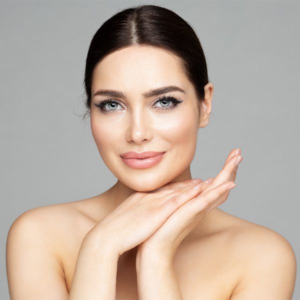 Woman with light skin, hands under chin, looking at camera. Light makeup, gray background.