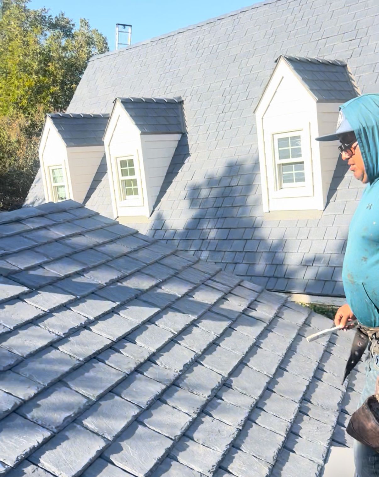Worker inspecting a gray shingled roof with dormer windows on a sunny day