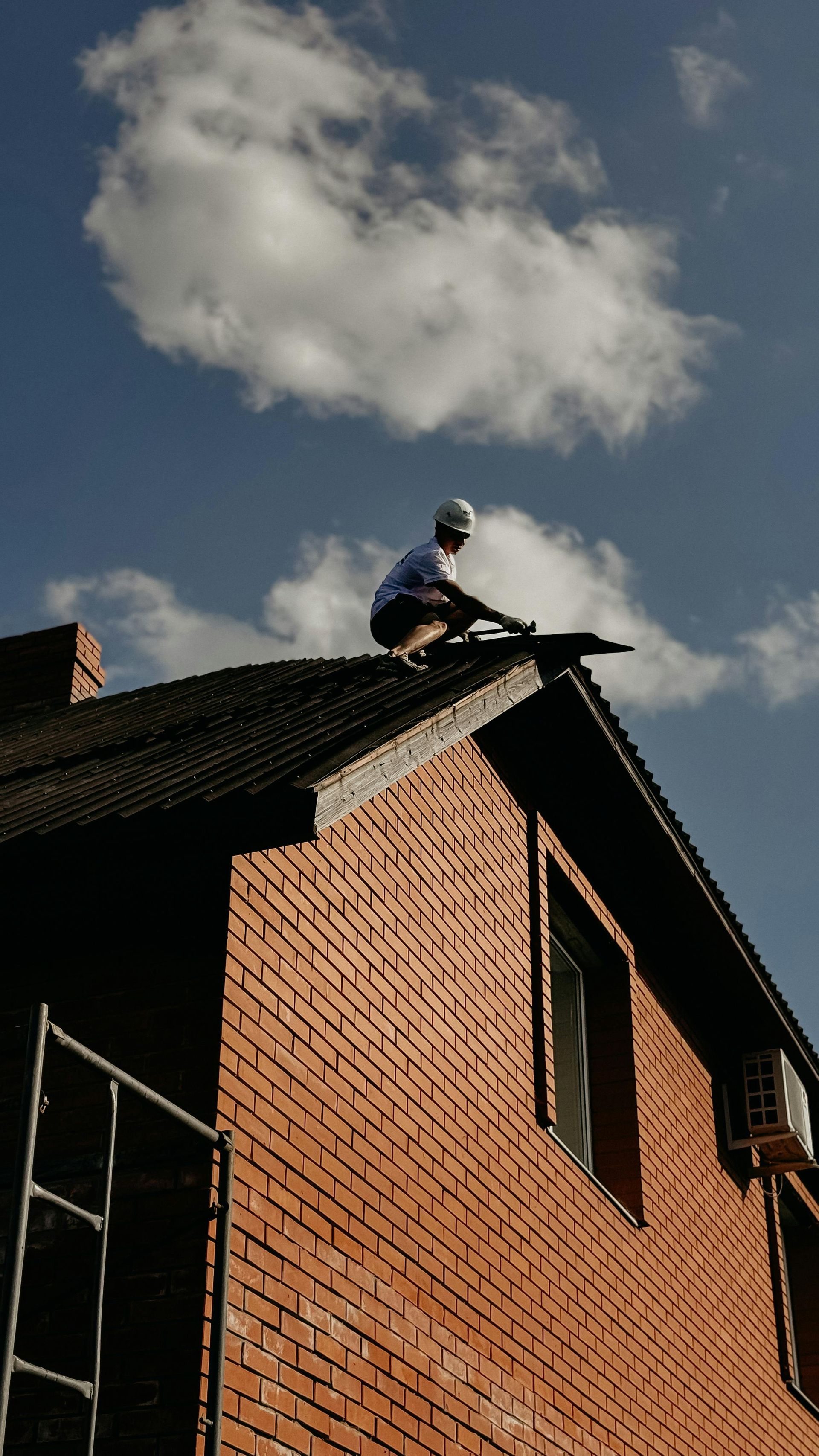 Worker on a brick house roof under a blue sky with clouds, next to scaffolding.