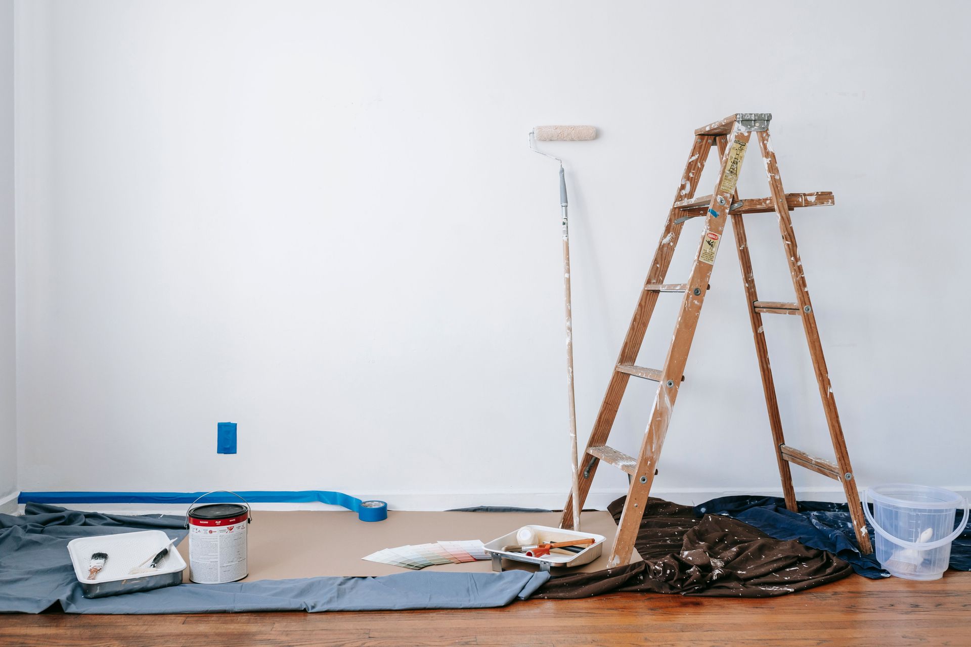 Paint supplies, roller, ladder, and drop cloth in an unfinished room with white walls and wood floor