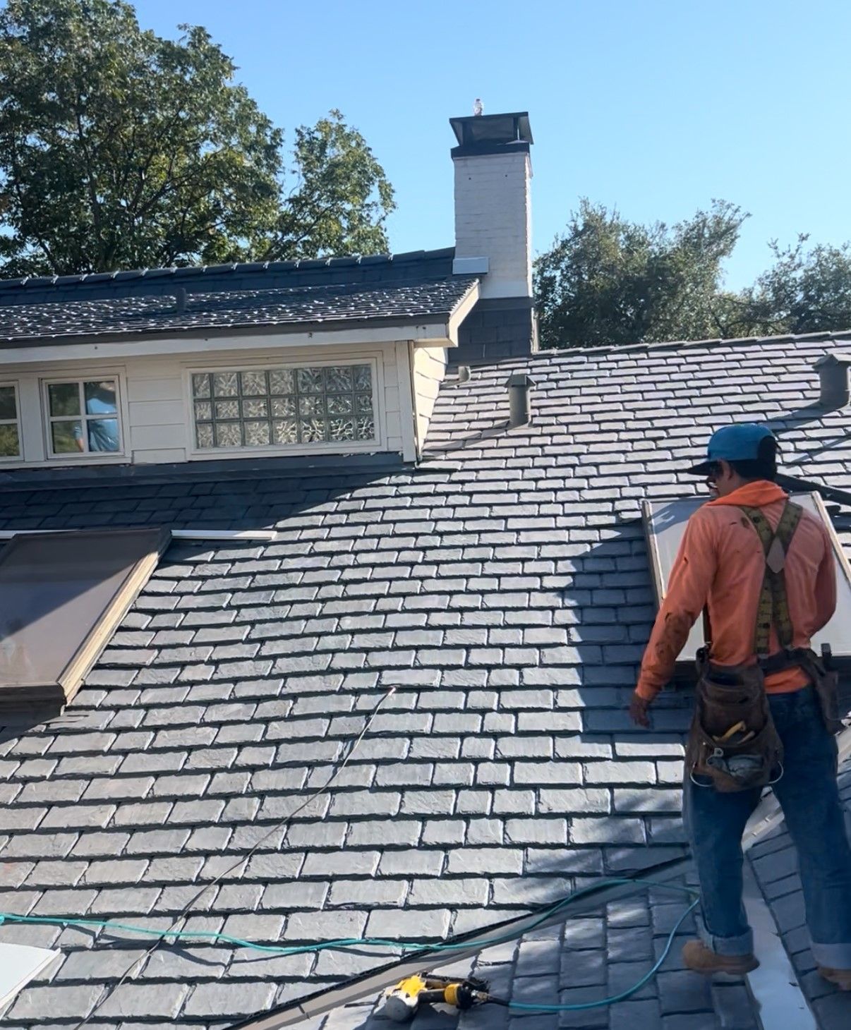 Roofer on a gray shingled roof near a chimney, working beside a house under a clear blue sky