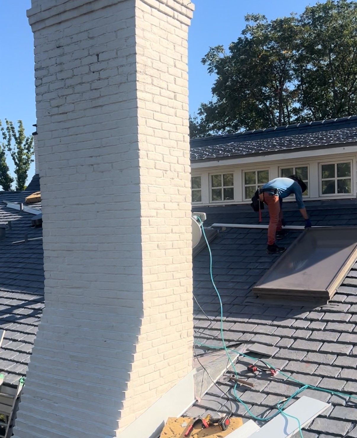 Worker repairing a gray shingled roof beside a white brick chimney on a sunny day