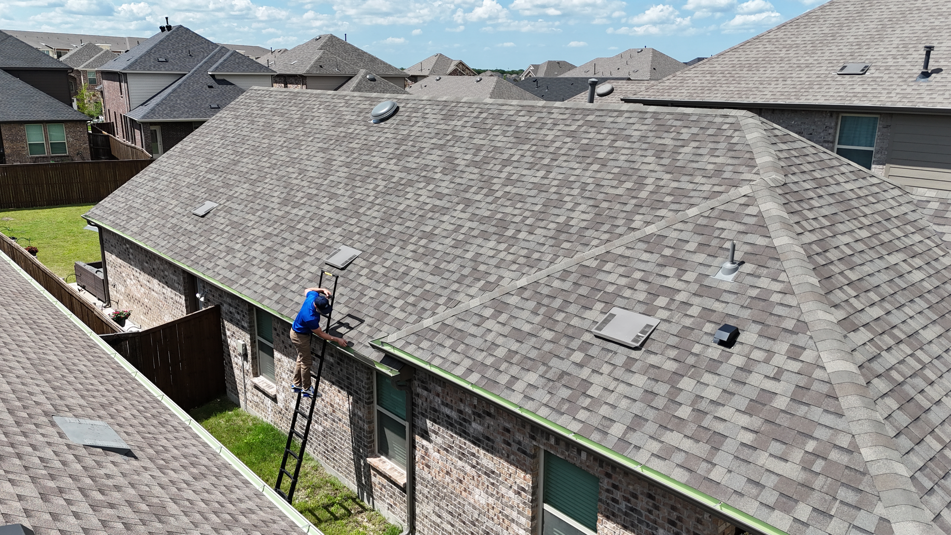 Worker on a ladder inspecting a gray shingled roof in a suburban neighborhood