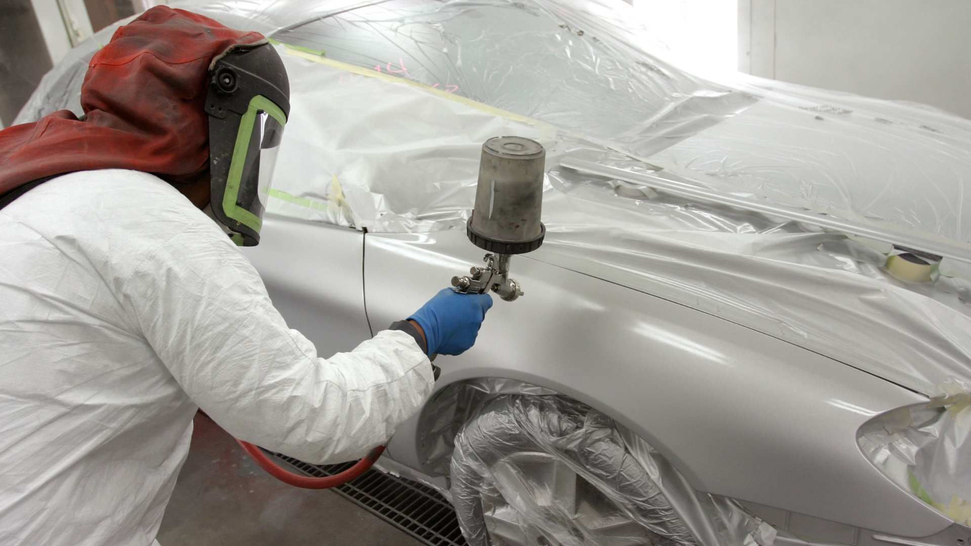 Worker spray-painting a car in a paint booth, wearing protective gear and a respirator