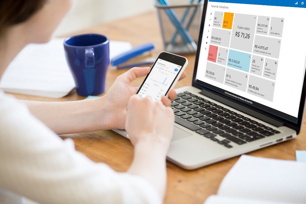 Uma mulher está sentada em uma mesa trabalhando pelo laptop e pelo celular.