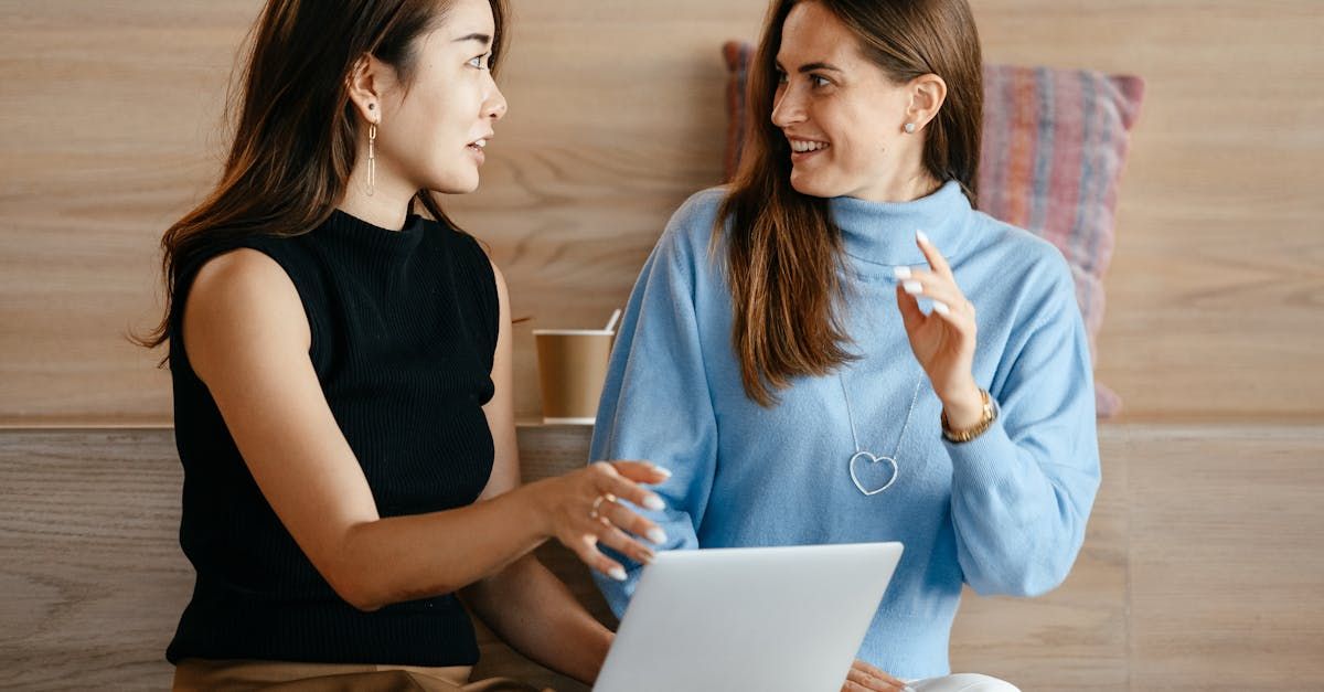 Duas mulheres estão sentadas à mesa com um laptop e conversando entre si