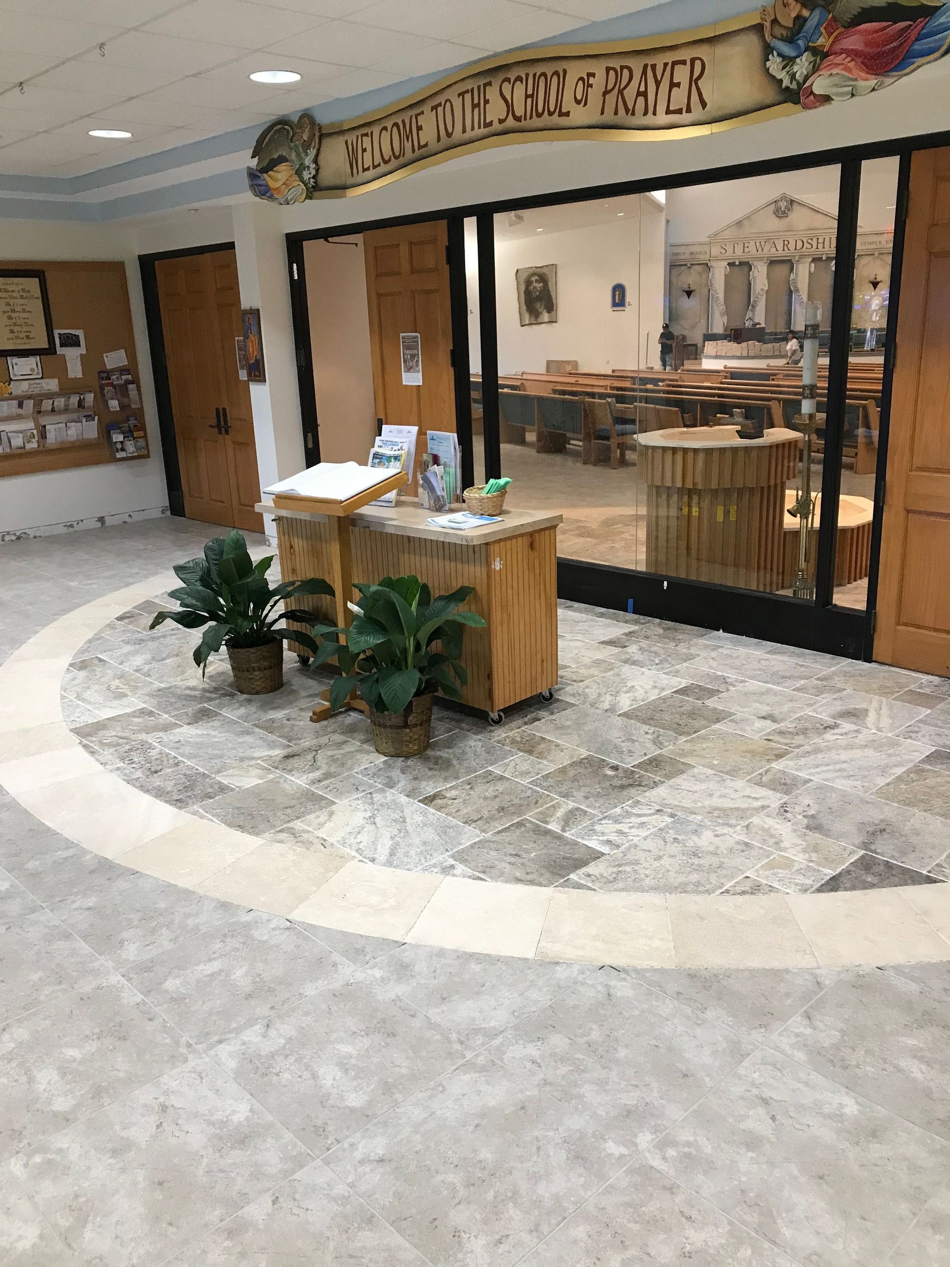 School lobby with a wooden reception desk, potted plants, and a glass-walled cafeteria area behind it