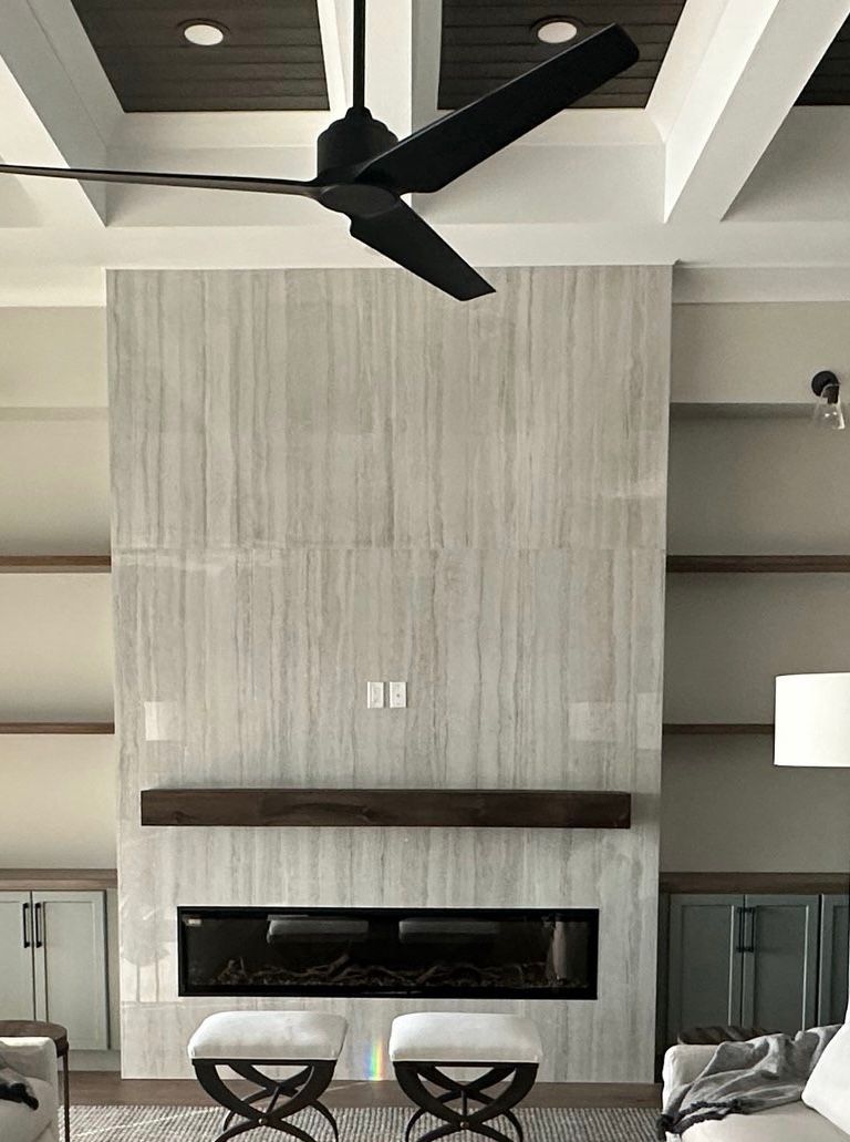 Modern living room with a tall fireplace, dark ceiling fan, and two round ottomans beneath a mantle