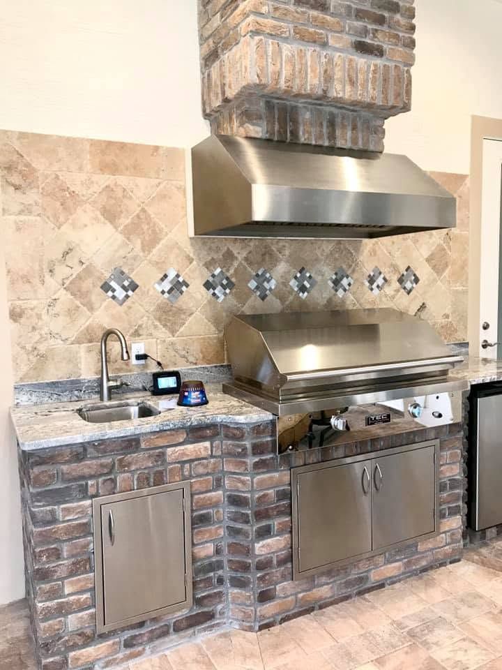 Stone outdoor kitchen with brick grill, stainless-steel hood, sink, and tiled backsplash