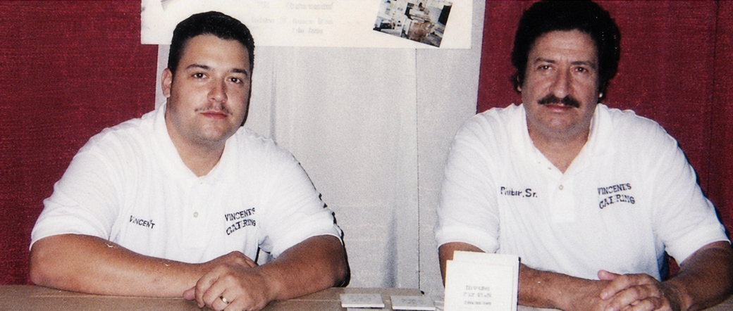 Two men in white shirts seated at a table, facing the camera in an indoor setting