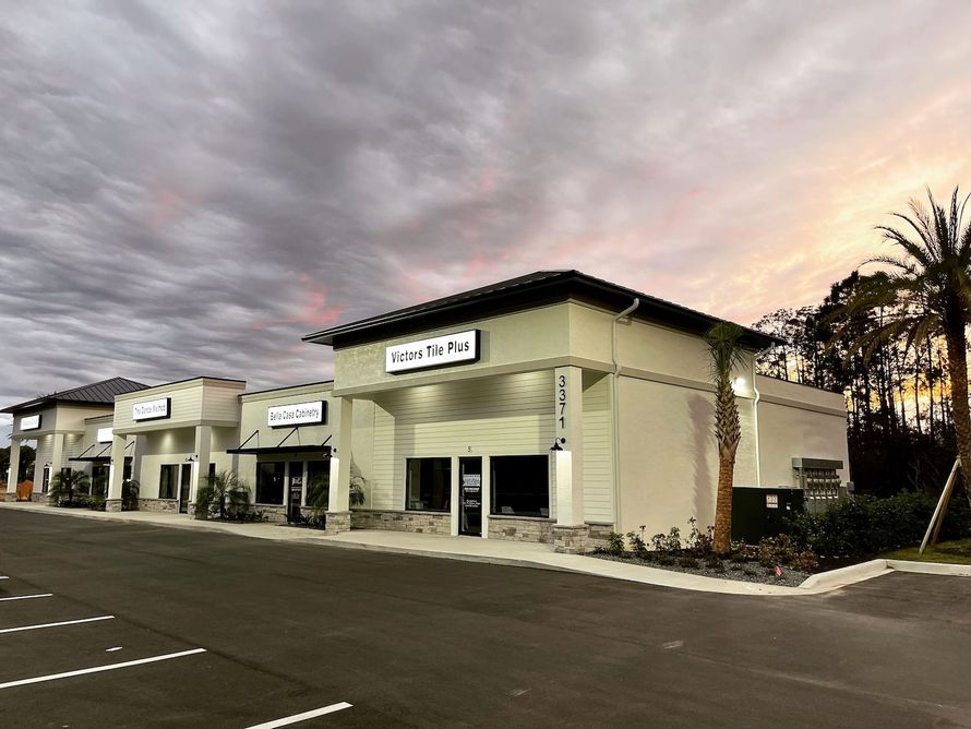 Strip mall storefronts at dusk under a cloudy sky, with palm trees and empty parking spaces.