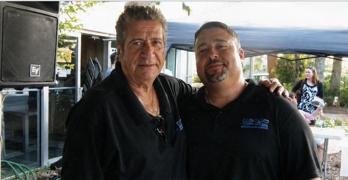 Two men standing together outdoors, one in a black shirt and the other in a black polo, smiling at the camera.