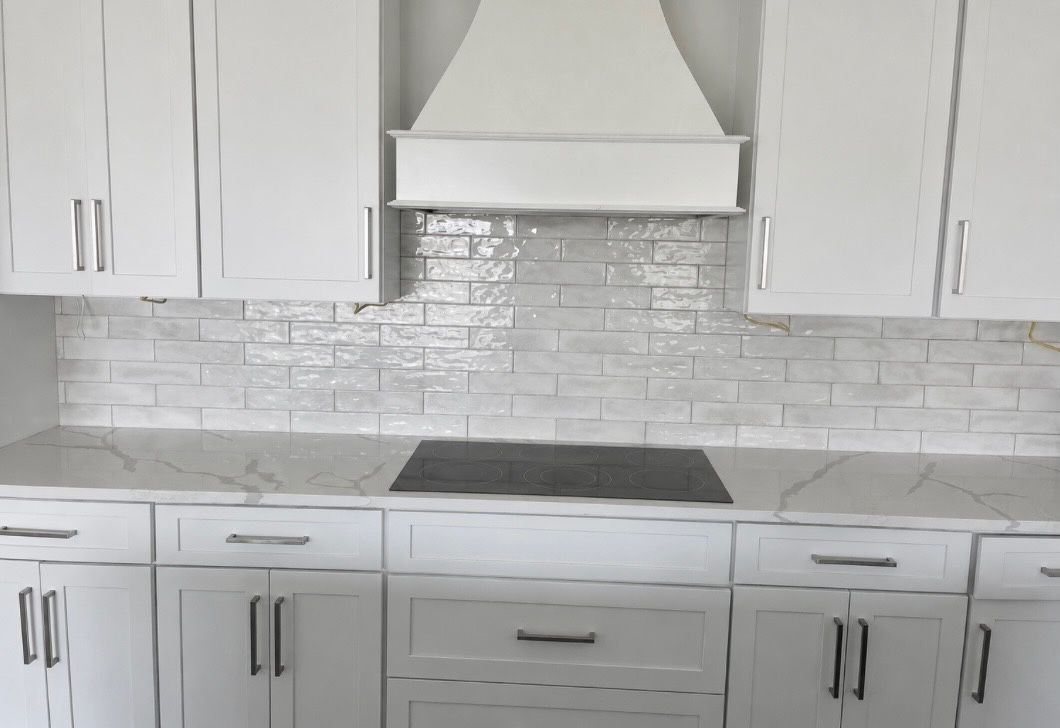 White modern kitchen with cabinets, white tile backsplash, and stainless range hood above cooktop