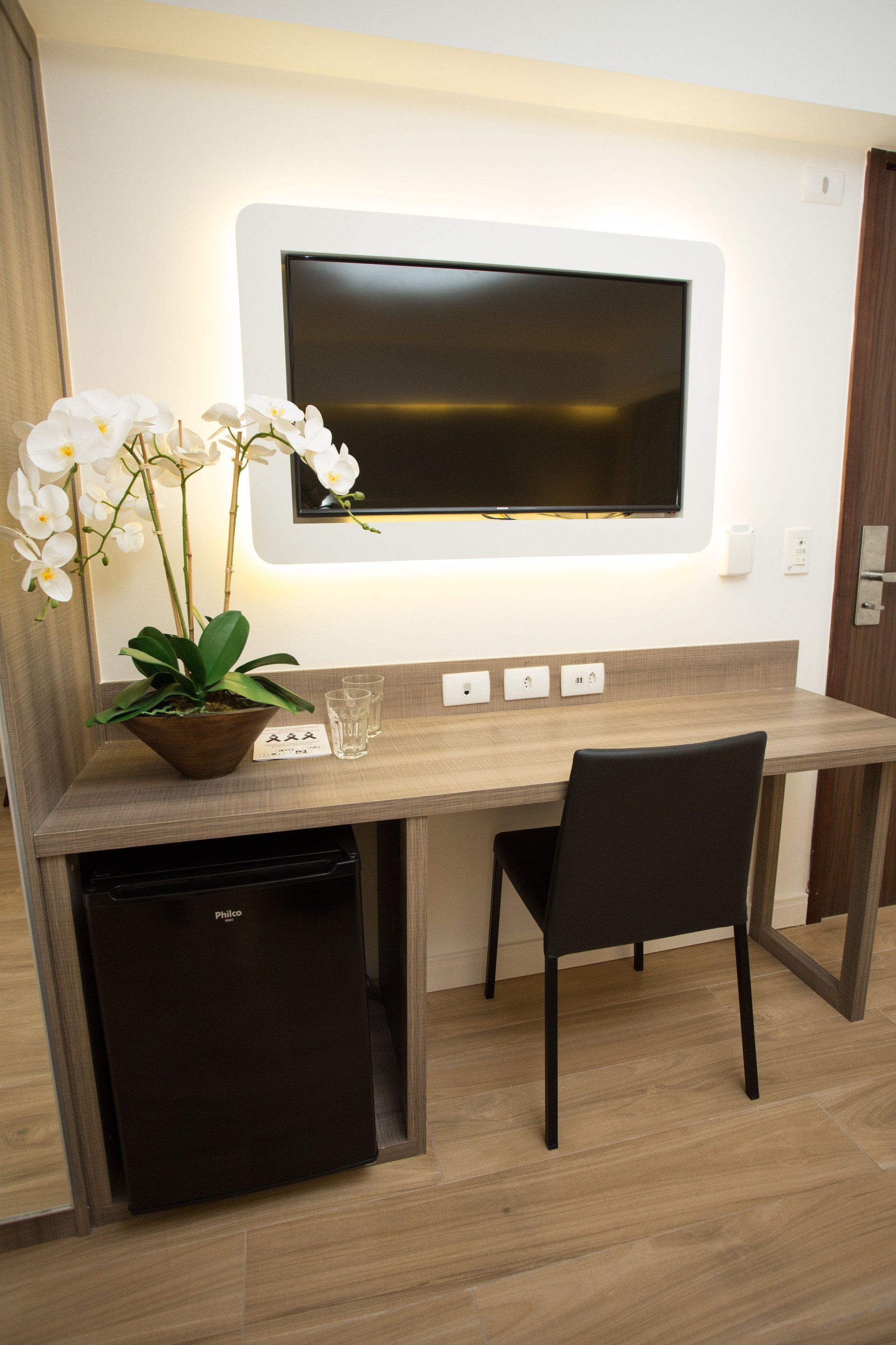 Um quarto de hotel com mesa, cadeira, televisão e geladeira.