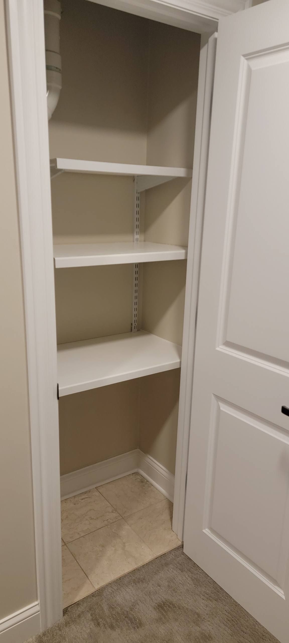 An open white closet door reveals three white shelves mounted on adjustable metal tracks against a light brown wall.