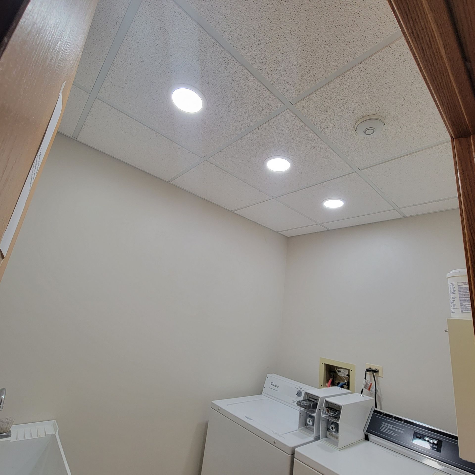 A small laundry room with white walls, a drop ceiling with three recessed lights, and two white laundry machines.