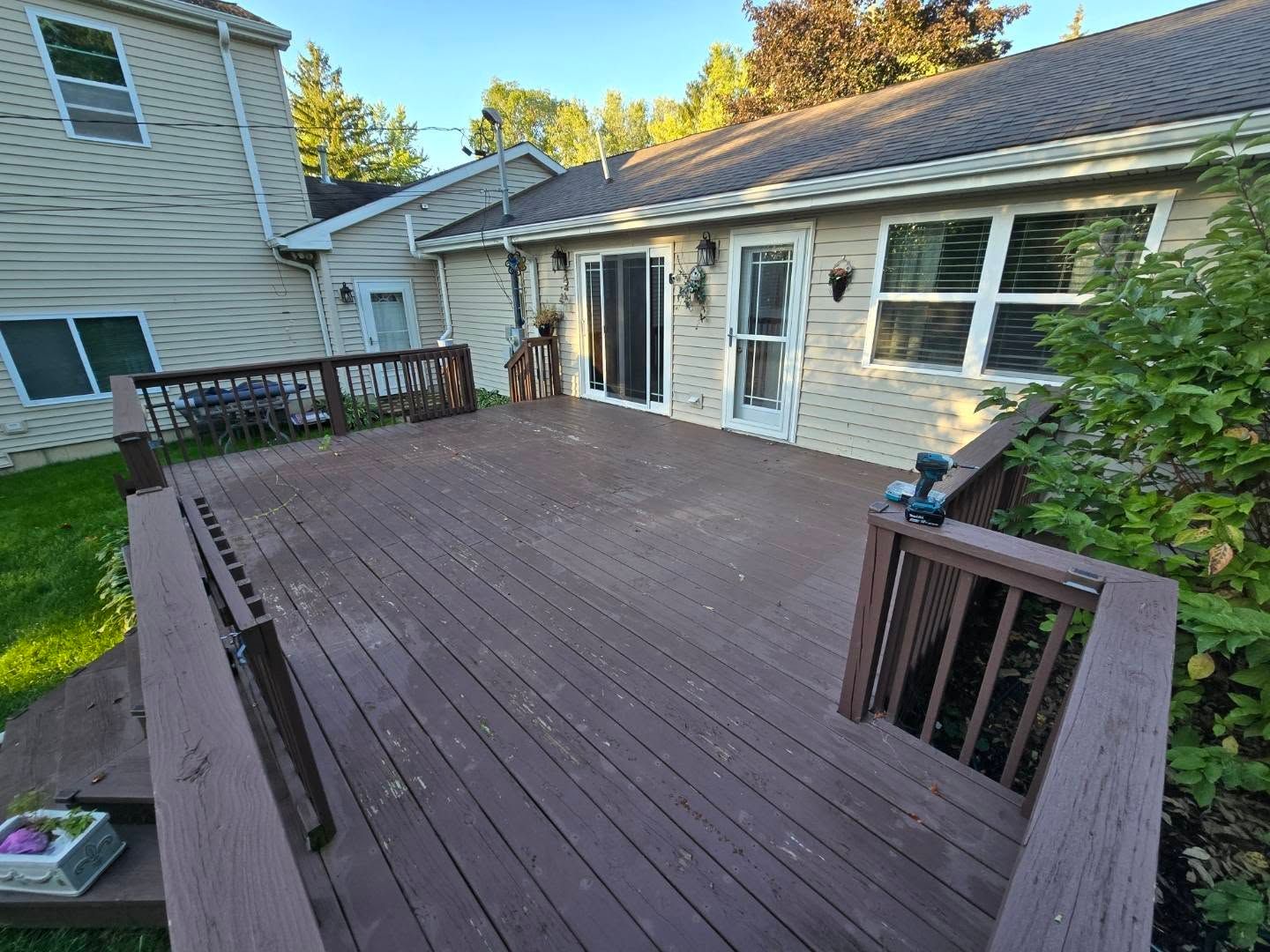 A wooden deck with brown railings extends from the back of a light-colored house with a sliding door and windows.