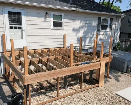 A wood deck frame under construction against the side of a light-colored house with a hot tub on the gravel ground.