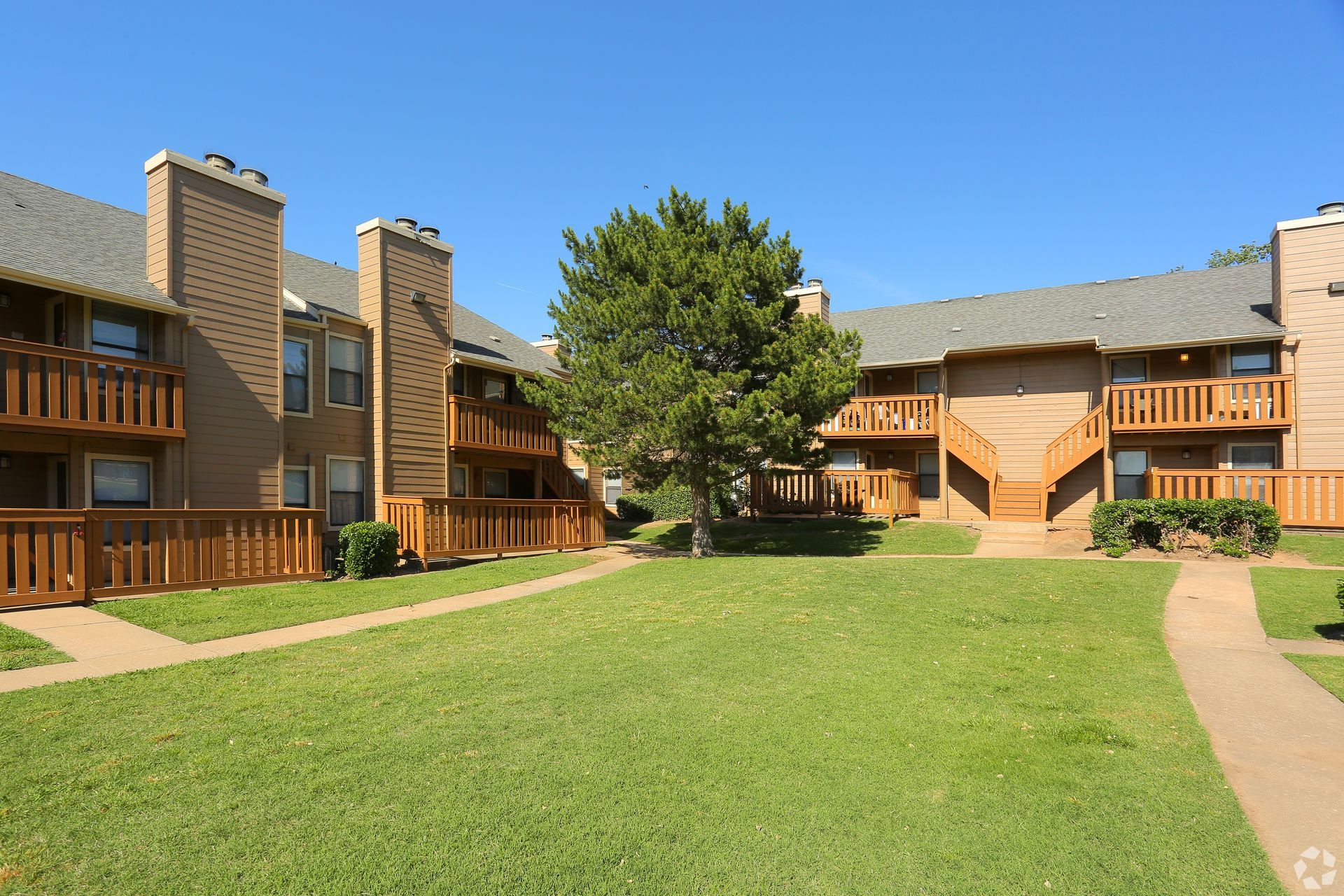Apartment courtyard with green lawn, trees, and tan brick buildings under a clear blue sky
