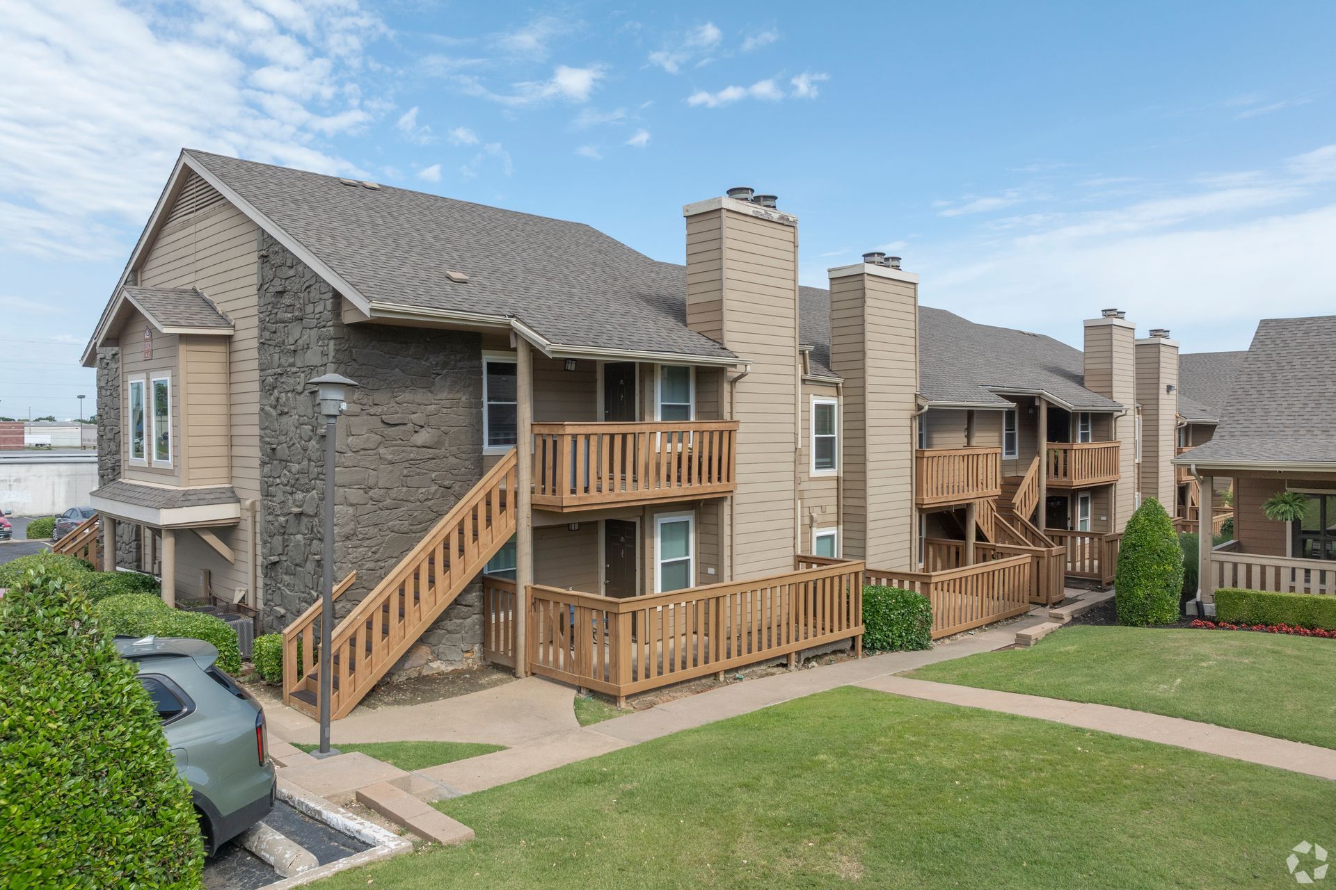 Apartment buildings with stone siding, balconies, stairs, and a parked car beside a grassy courtyard