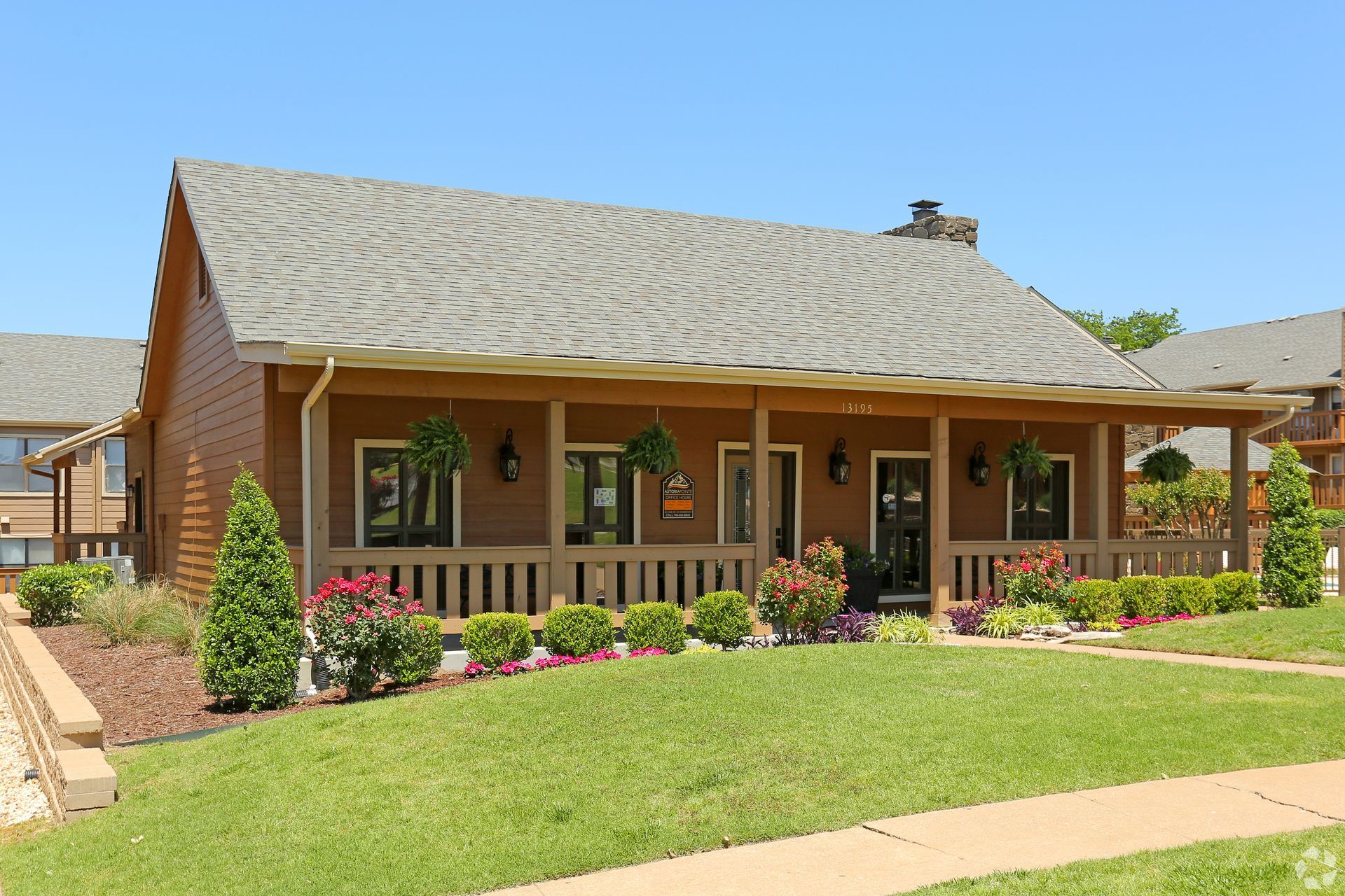 Single-story wooden house with a porch, green lawn, and flower beds under a clear blue sky