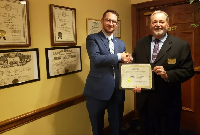 Two men in suits shake hands while holding a framed certificate in an office hallway.