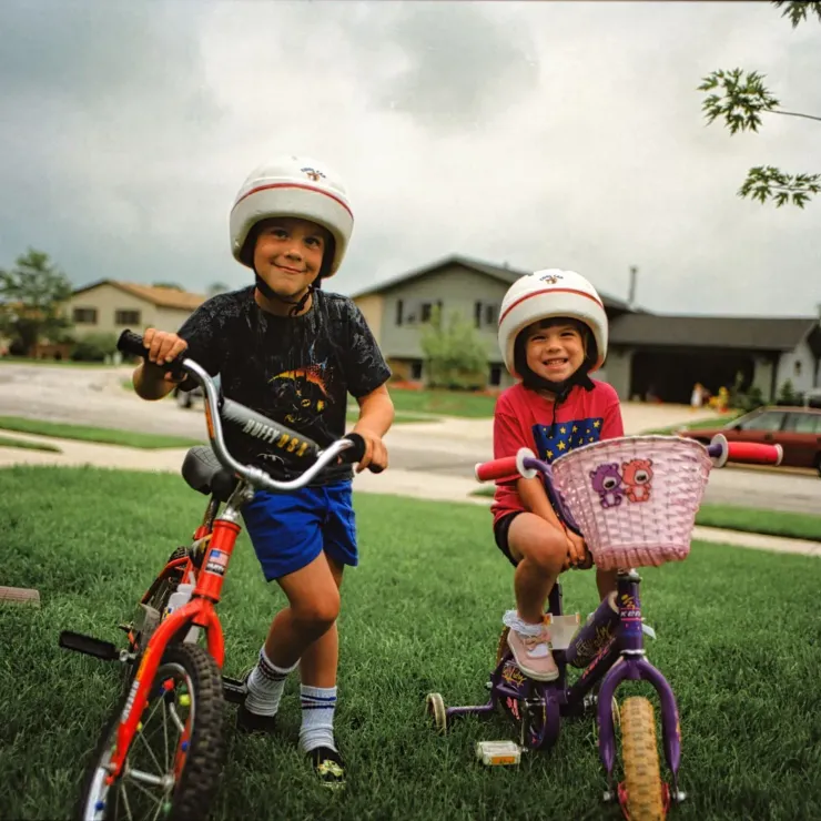 Two children in helmets ride bikes on a suburban street, smiling near houses and green lawns.