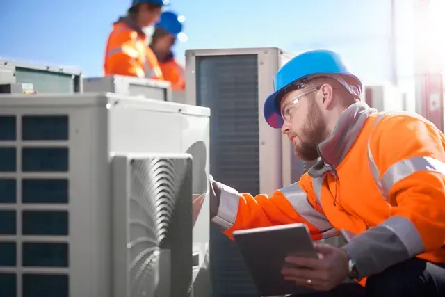 Technician in orange safety gear inspecting outdoor HVAC unit with tablet and clipboard