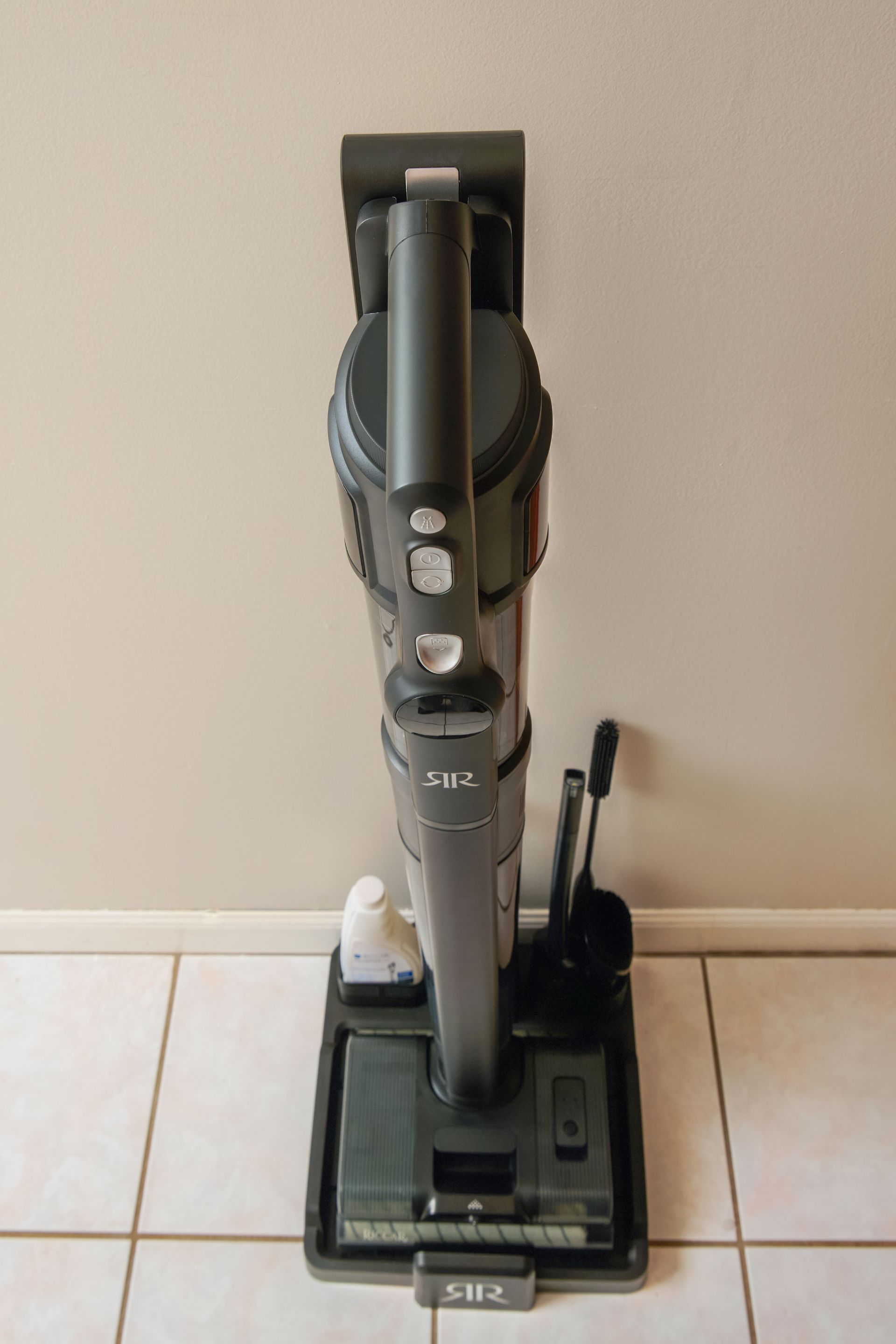Upright dark gray vacuum cleaner against a beige wall on tiled floor.