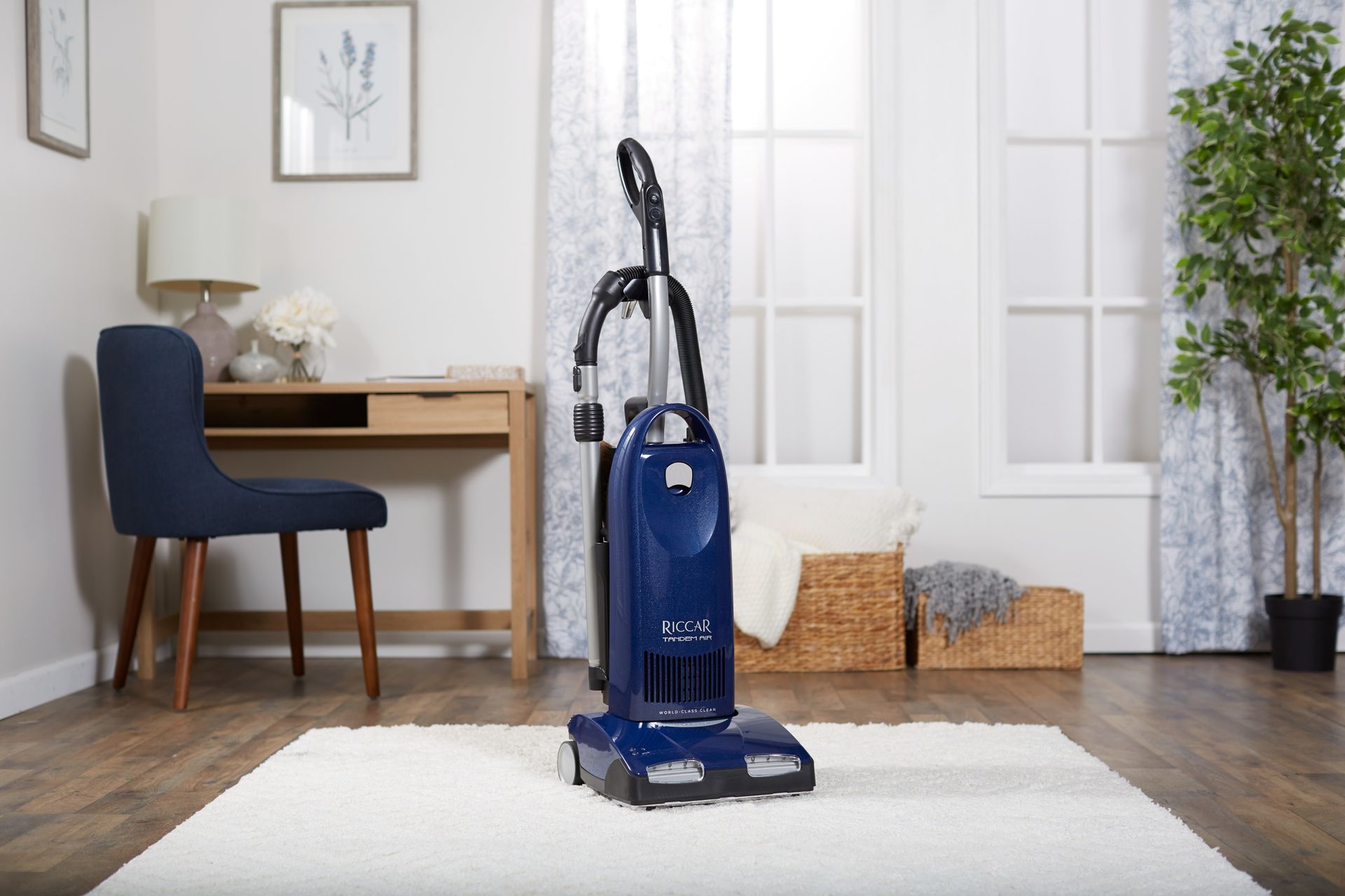 Blue upright vacuum cleaner on a white rug in a room with a desk, chair, and window.