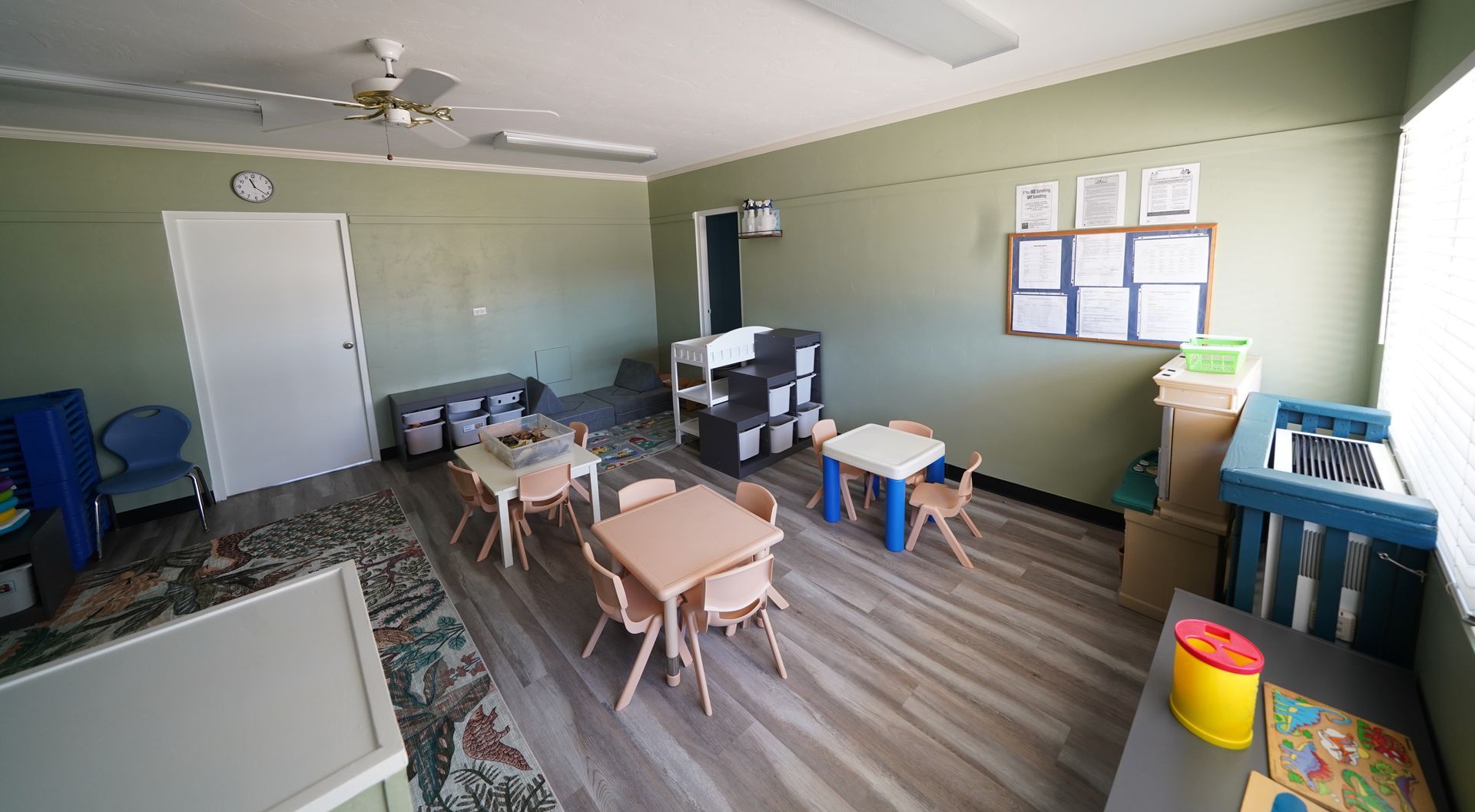 Classroom with small tables, chairs, whiteboard, bulletin board, and toy bins on a wood floor.