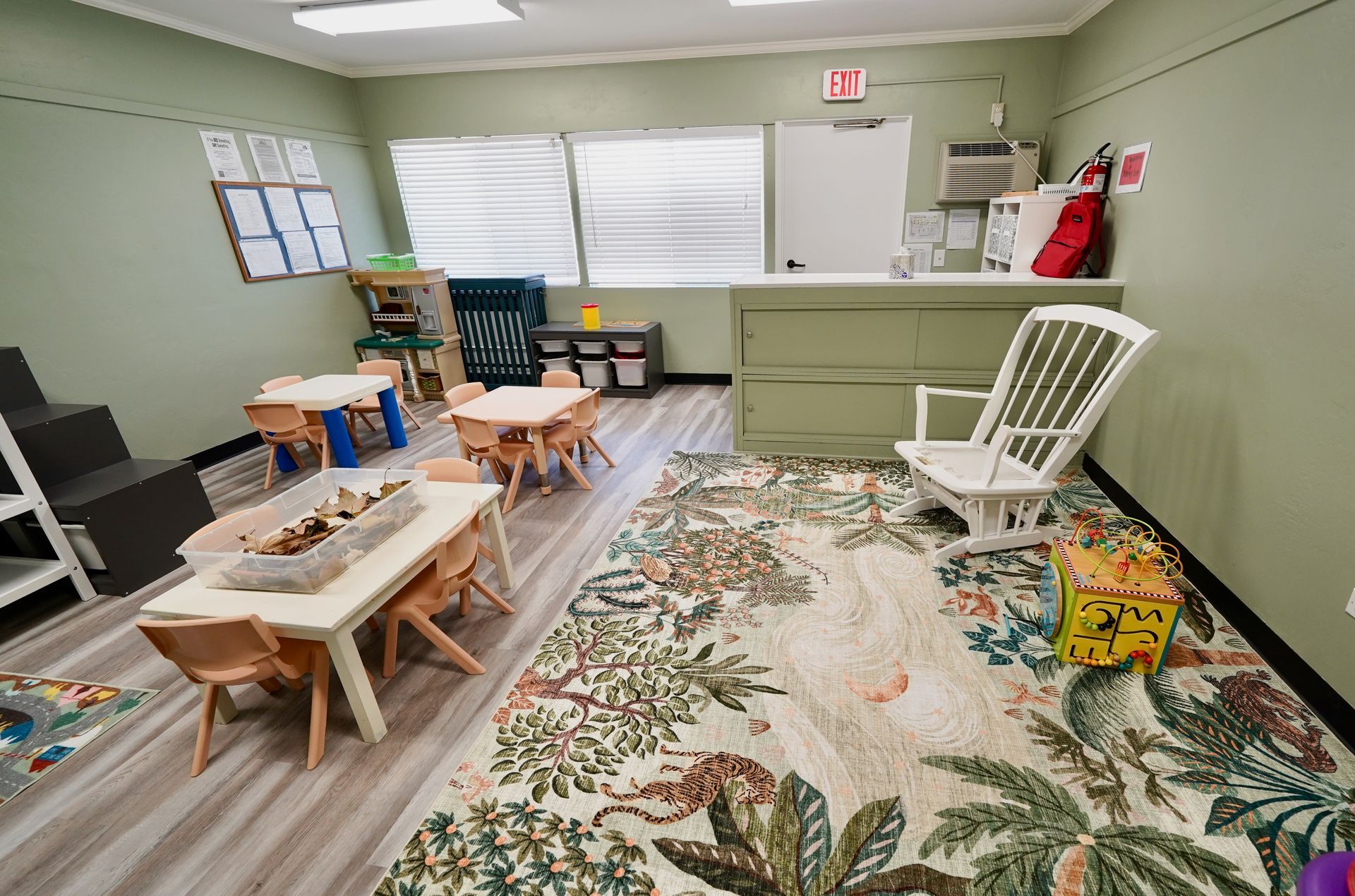 Playroom with low tables, toys, a patterned rug, green walls, and a white rocking chair by the window.