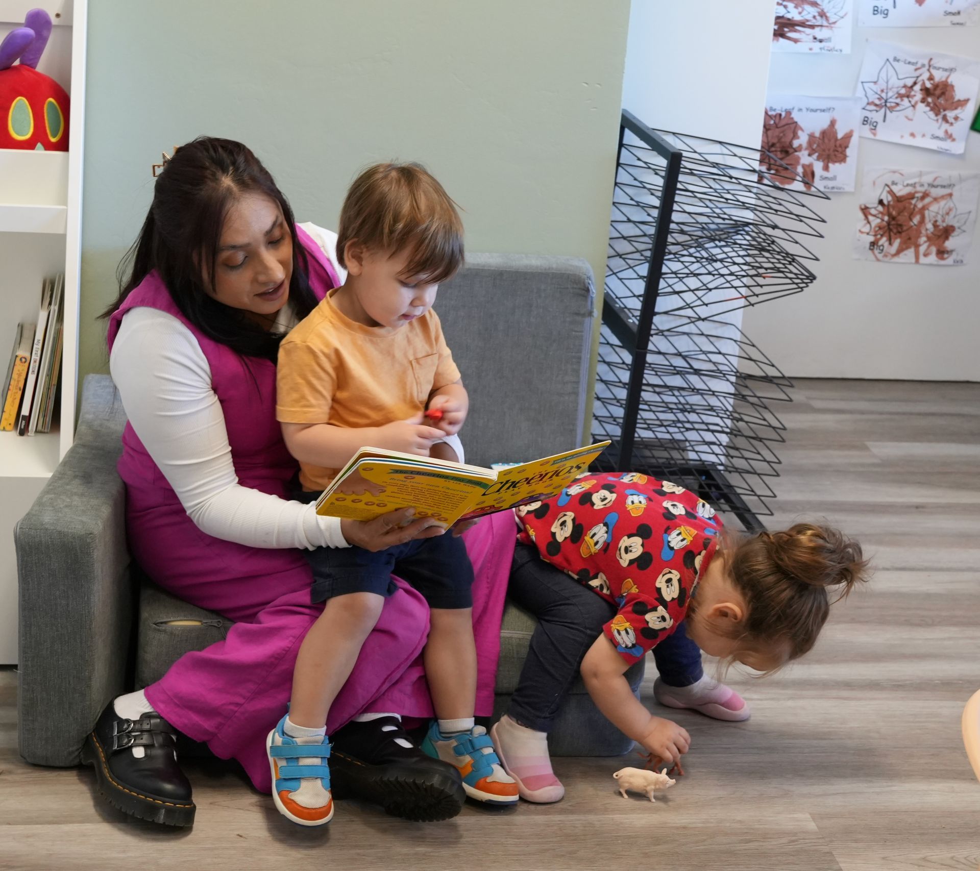 Adult reading with two children in a waiting room, while another child crouches on the floor nearby.