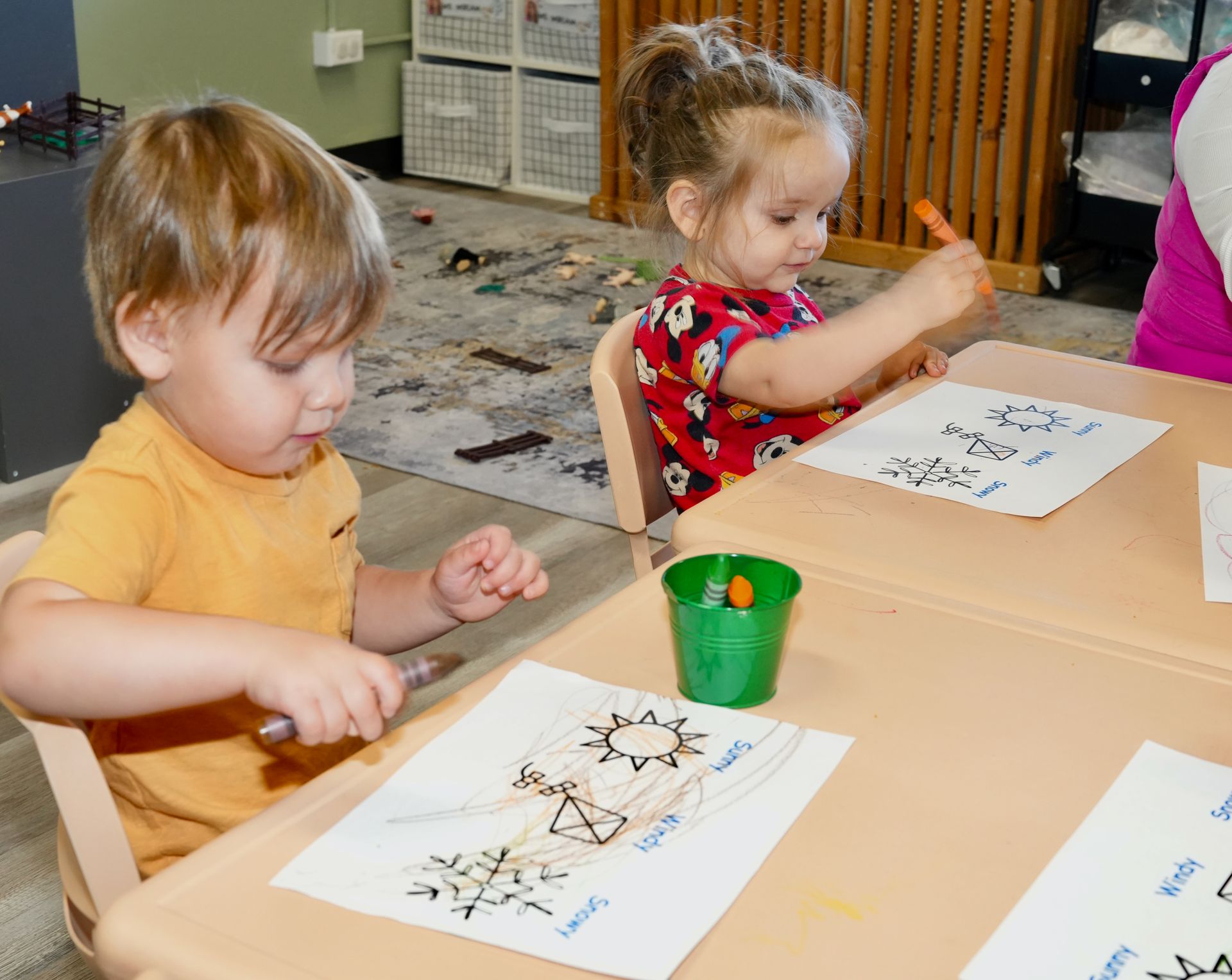 Two children painting at a table with paper and cups of paint in a classroom