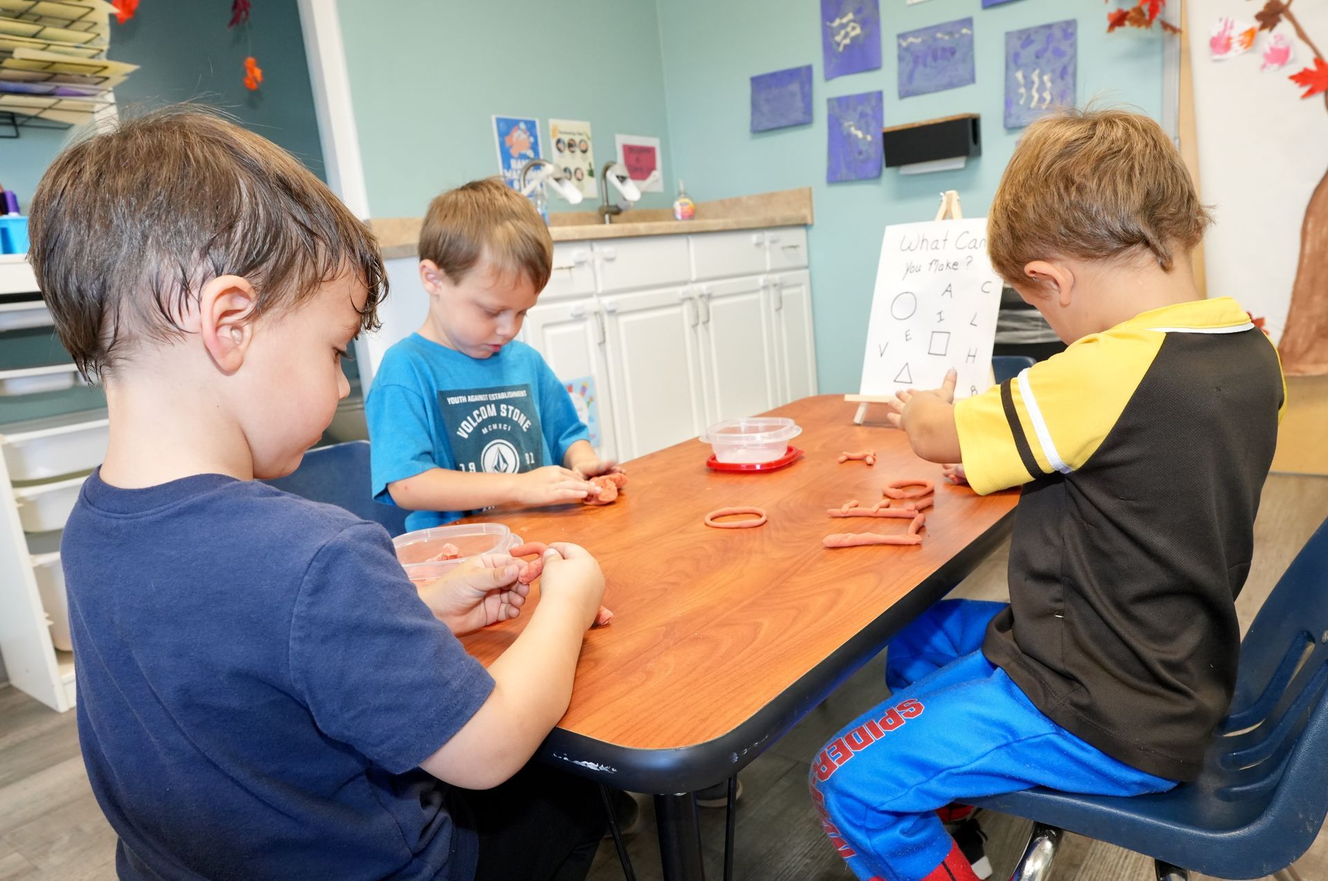 Three children seated at a table, playing with clay in a classroom.