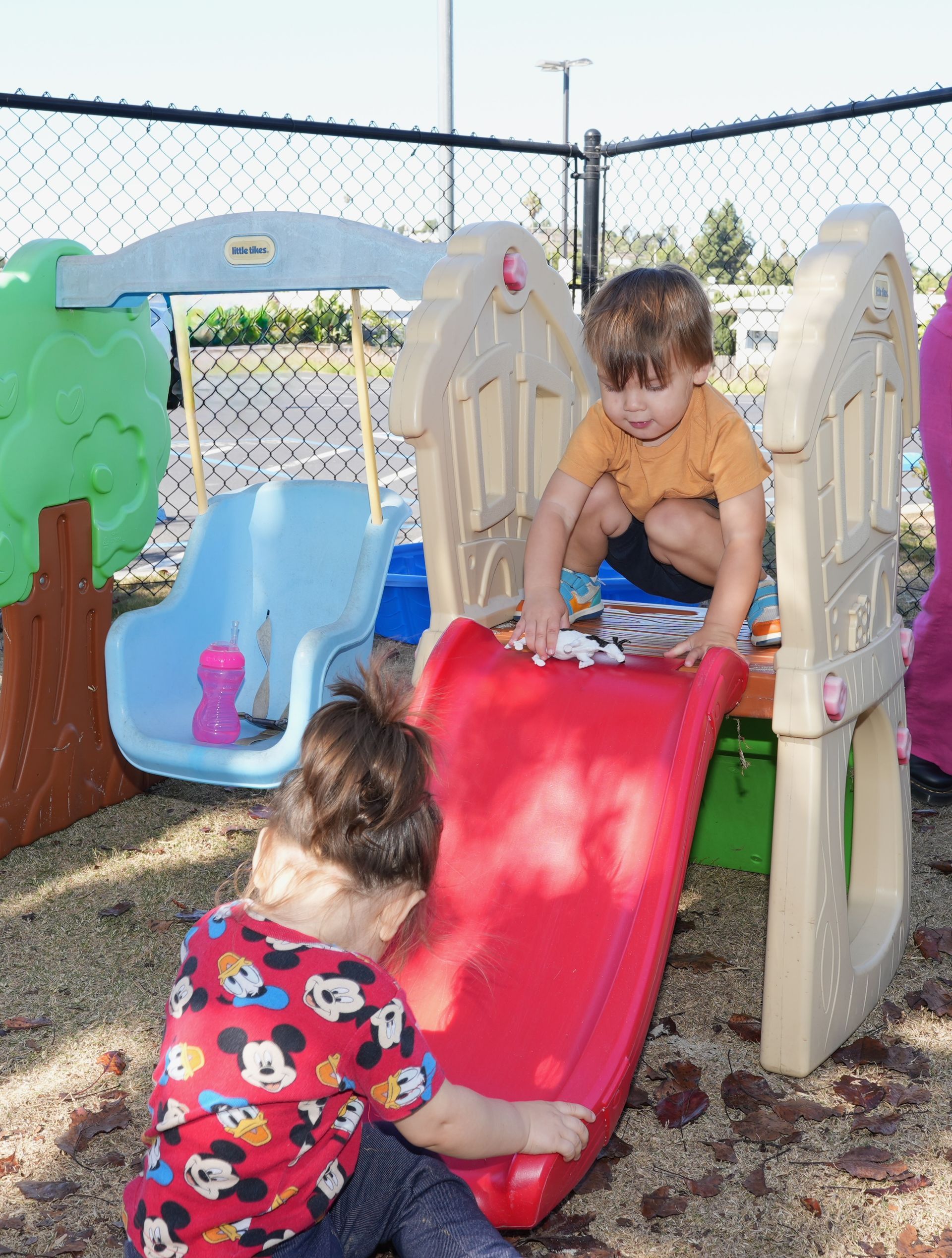Two children playing on a red slide and beige play structure in a fenced playground