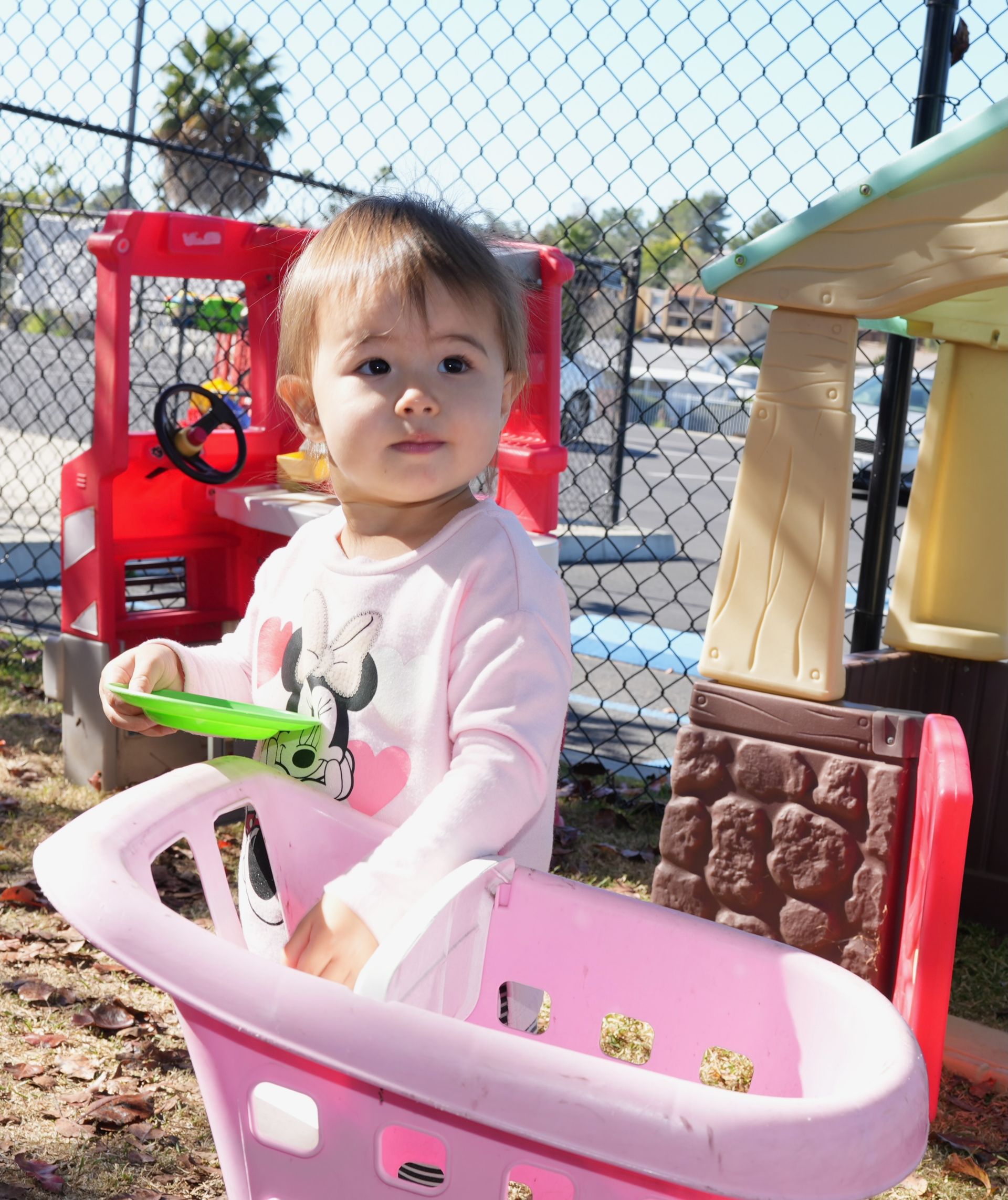 Toddler in a pink shirt sitting in a pink toy cart outdoors near a fence and play equipment