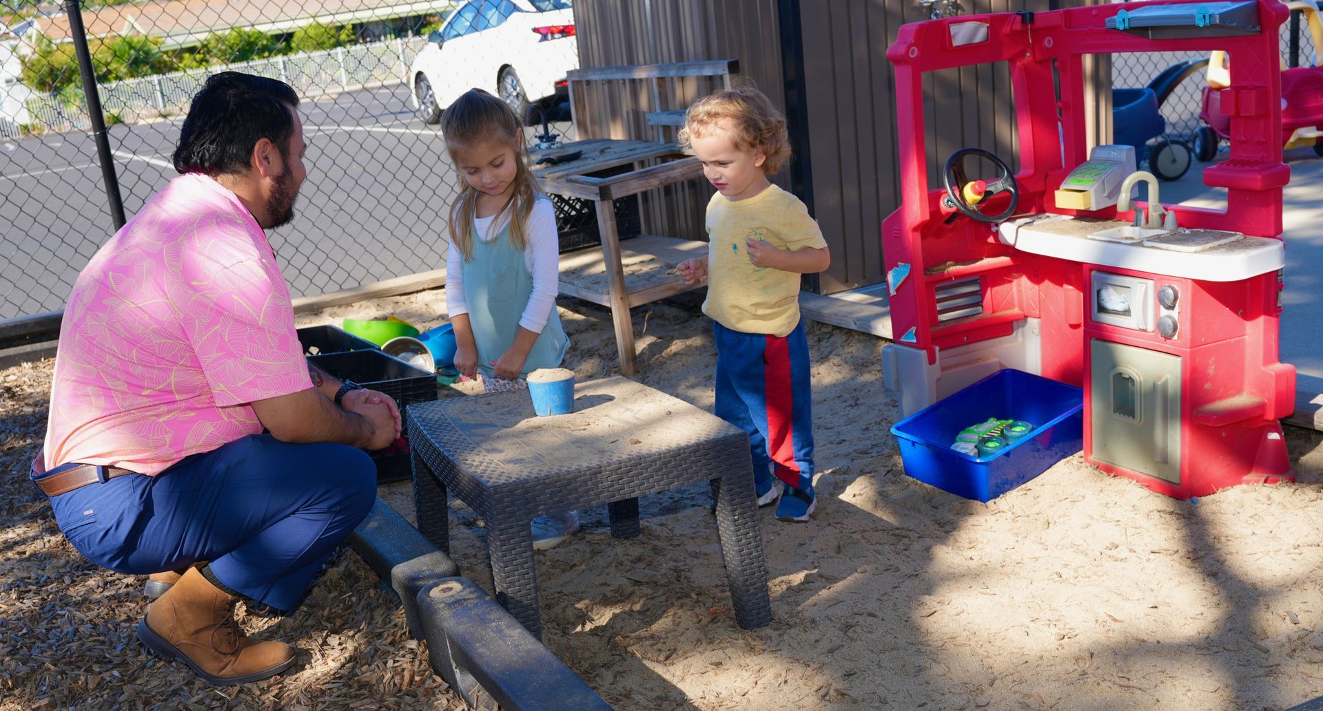 People sitting around a picnic table outdoors beside a red play kitchen, chatting in a sandy yard.