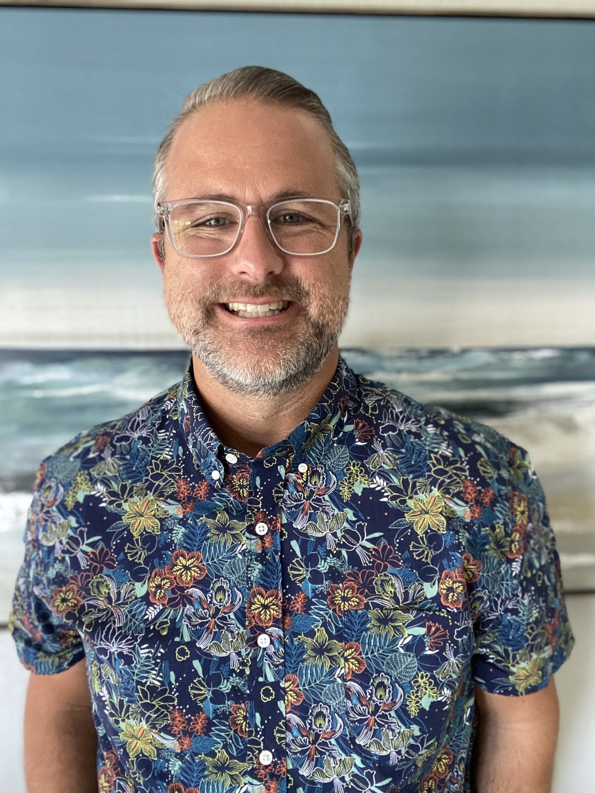 Smiling person in glasses wearing a blue floral shirt against a blurred ocean backdrop