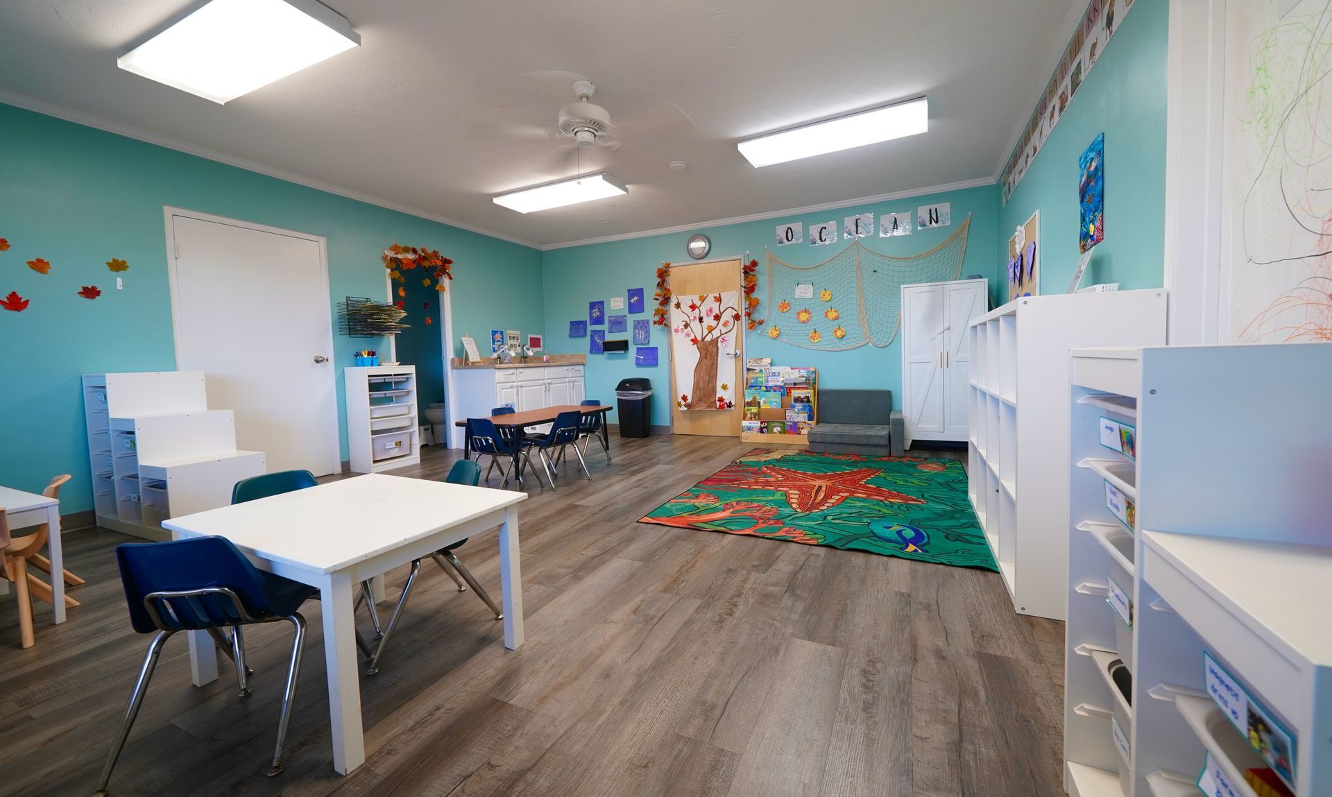 Bright classroom with tables, chairs, cubbies, colorful rug, and children’s artwork on teal walls.