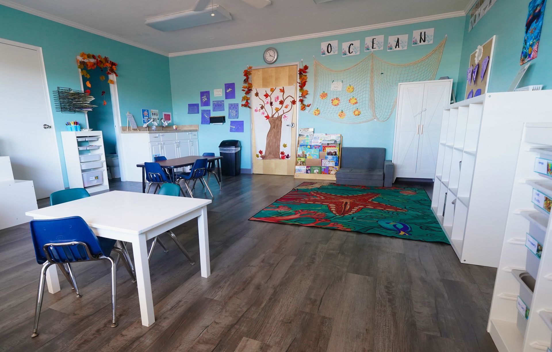 Empty classroom with teal walls, white desks, blue chairs, colorful wall art, and a red rug on wood floor