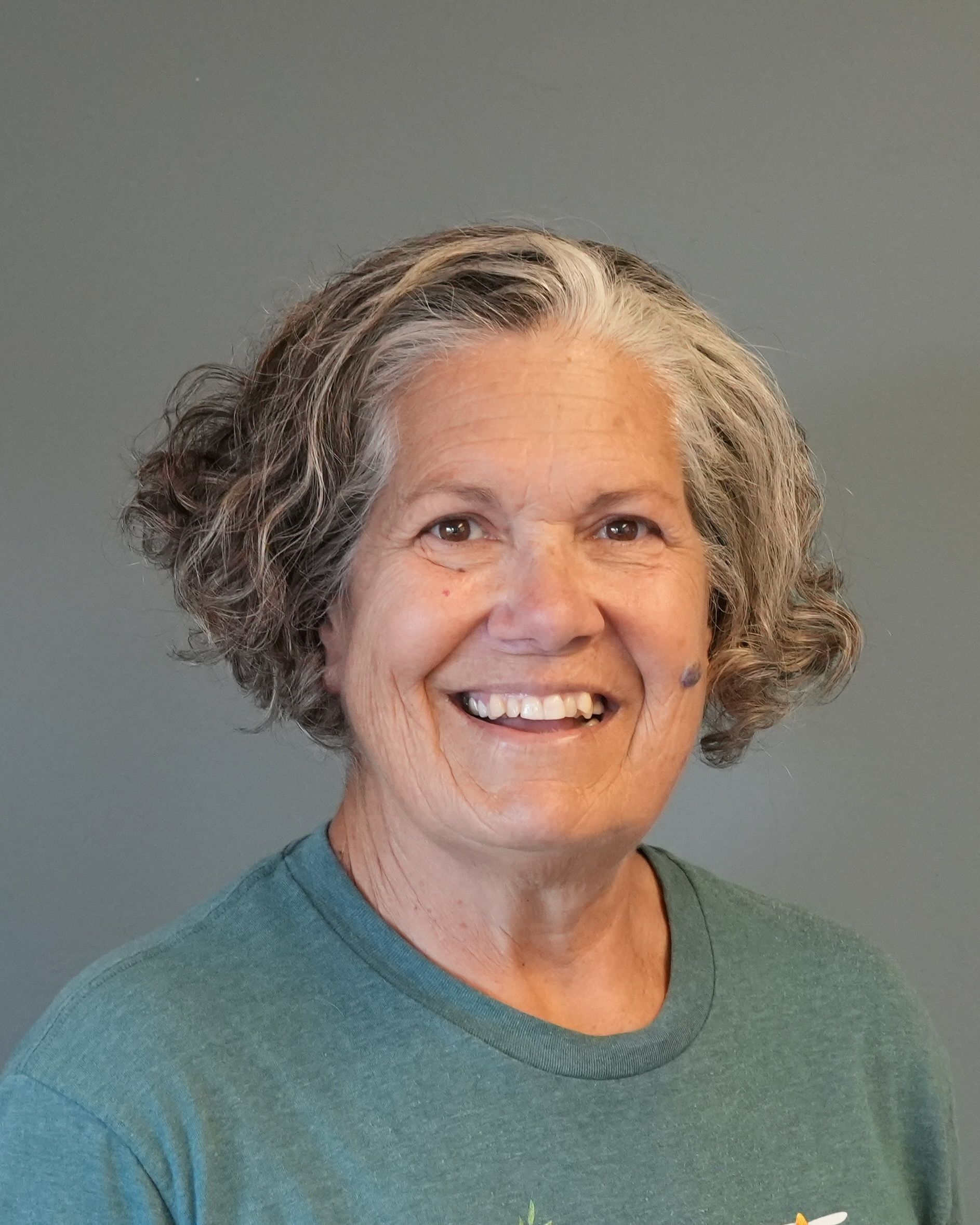 Smiling person with short gray hair wearing a teal shirt against a gray background